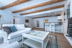 Ein modernes Wohnzimmer mit einem weißen Sofa, einem Glastisch, Holzbalken an der Decke, einer Küchenzeile mit weißen Barhocker und einem großen Fenster.
