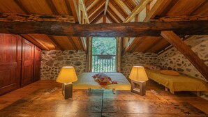Un dormitorio en el ático con pared de piedra, vigas de madera, una cama con manta, dos lámparas de noche y una ventana con vistas a la vegetación.