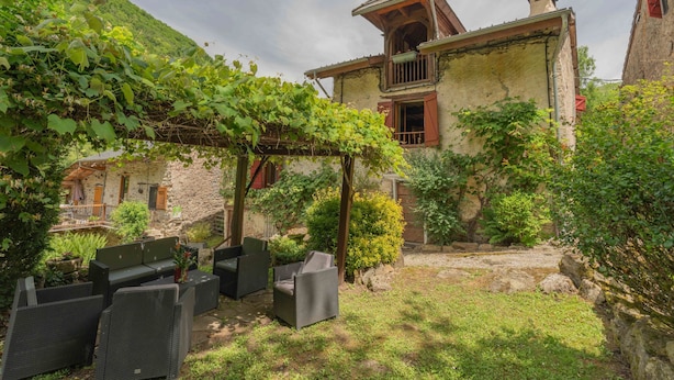 Un edificio rústico de piedra con un balcón, una pérgola cubierta de hiedra y mobiliario de exterior.