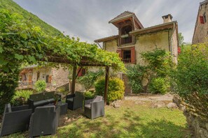 A rustic stone building with a balcony, a pergola with vines, and outdoor seating.