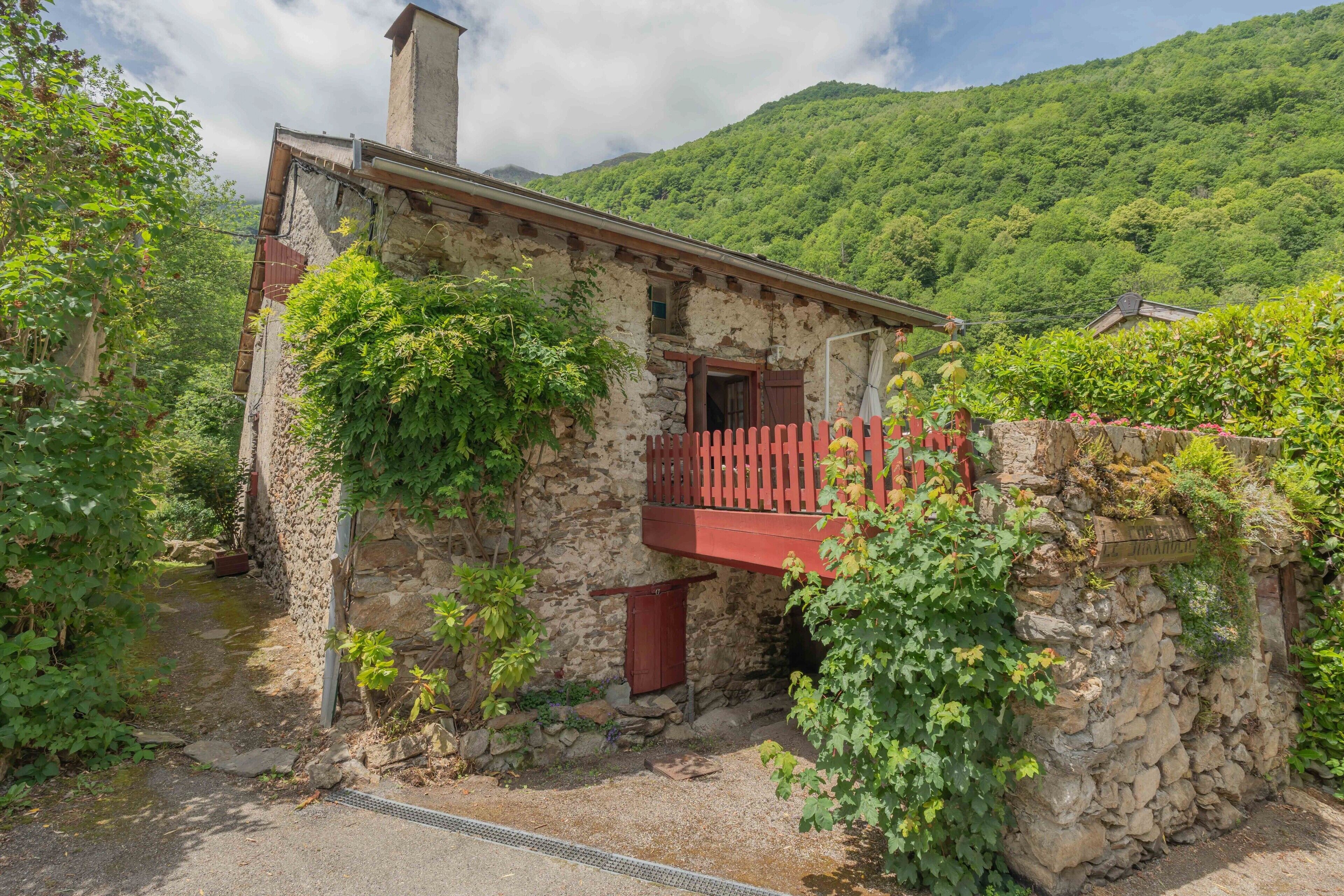 Un edificio de piedra con un balcón rojo, rodeado de follaje y con una montaña al fondo.