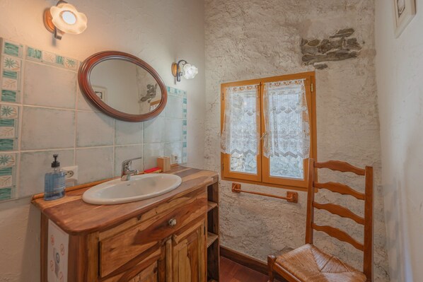A bathroom with a wooden vanity, a round mirror, a sink, a window with lace curtains, and a wooden chair.