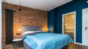 A bedroom with a brick wall, a bed with a blue quilt, a round side table, and a door leading to another room. - Designer Two-Bedroom Loft with Terrace in Old Town (Praha 1)