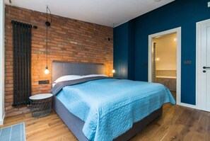 Ein Schlafzimmer mit Backsteinwand, einem Bett mit blauer Steudecke, einem runden Beistelltisch und einer Tür zu einem anderen Raum.