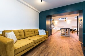 Ein modernes Wohnzimmer mit einem gelben Sofa, einem Essbereich mit Tisch und Stühlen sowie einer durchgehenden Tür einsehbarer Küche.