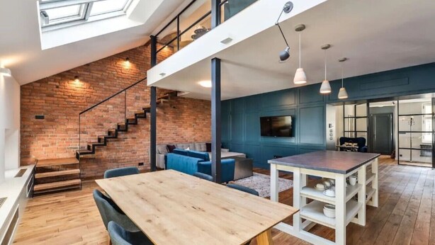 A loft-style room with a wooden dining table, a staircase, and a blue accent wall. - Designer Two-Bedroom Loft with Terrace in Old Town (Praha 1)