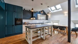 A modern kitchen with dark blue cabinets, a central island, and a dining area with a high ceiling and skylights. - Designer Two-Bedroom Loft with Terrace in Old Town (Praha 1)