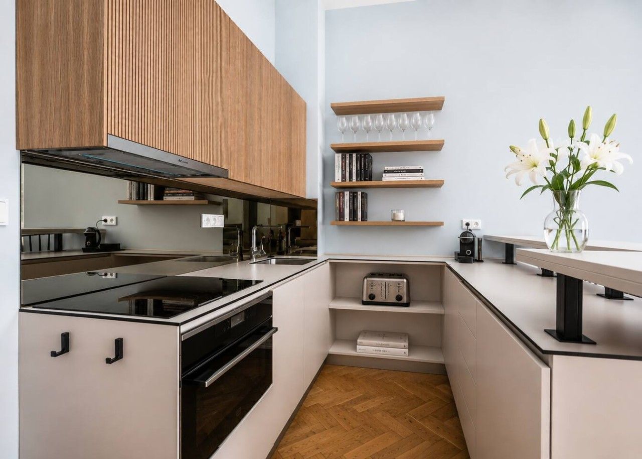 A modern kitchen with a built-in oven, a stovetop, and wooden cabinets. There are shelves with books and a vase with flowers.