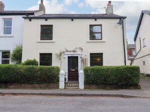 A two-story building with a white facade, a dark brown door, and a small garden area with neatly trimmed hedges.