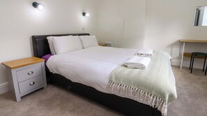 A neatly made bed with white linens, a bedside table with drawers, a small desk with a chair, and two wall-mounted light fixtures. - 6 Hazel Moor (Blackburn)