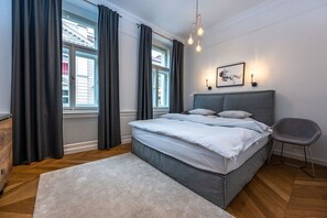 Ein Schlafzimmer mit einem Bett, einem Sessel, einem Teppich, einem Fenster mit Vorhängen und einer Wandleuchte.