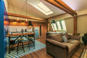Ein gemütlicher Wohnbereich mit Sofa, Esstisch und Hocker, einer Küche mit Backsteinwand und einem Oberlicht.