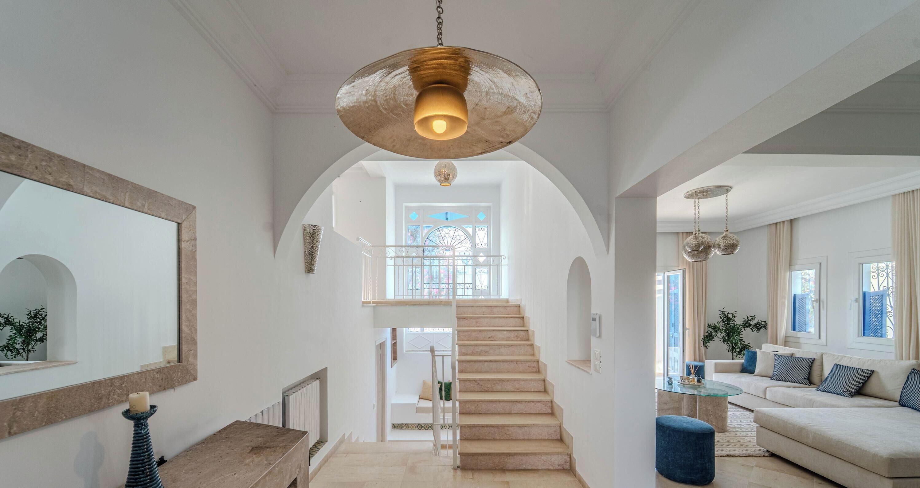 A spacious living area with a staircase leading to a balcony, a large mirror, and a hanging light fixture.