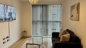 Una sala de estar moderna con televisor, sofá, una mesa de centro de vidrio, candelabro y un amplio ventanal con persianas.