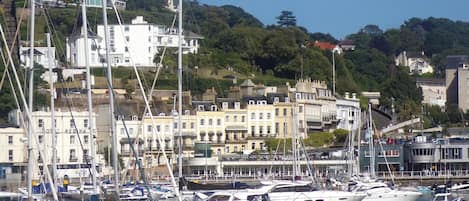 A marina with multiple boats docked, surrounded by buildings with a hilly backdrop.