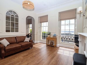 Ein geräumiges Wohnzimmer mit braunem Ledersofa, Holzboden, großen Fenstern mit Aussicht und einem Holztisch mit einer Vase und Büchern.