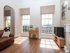 A living room with a brown sofa, wooden TV stand, and a large window with a view of a balcony and cityscape. - Flat 2 The Moorings (Torquay)