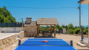 Uma mesa de bilhar com superfície azul e bochas vermelhas, cercada por paredes de pedra e uma pérgola.