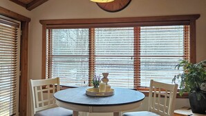 A dining area with a round table, four chairs, a clock on the wall, and large windows with blinds.