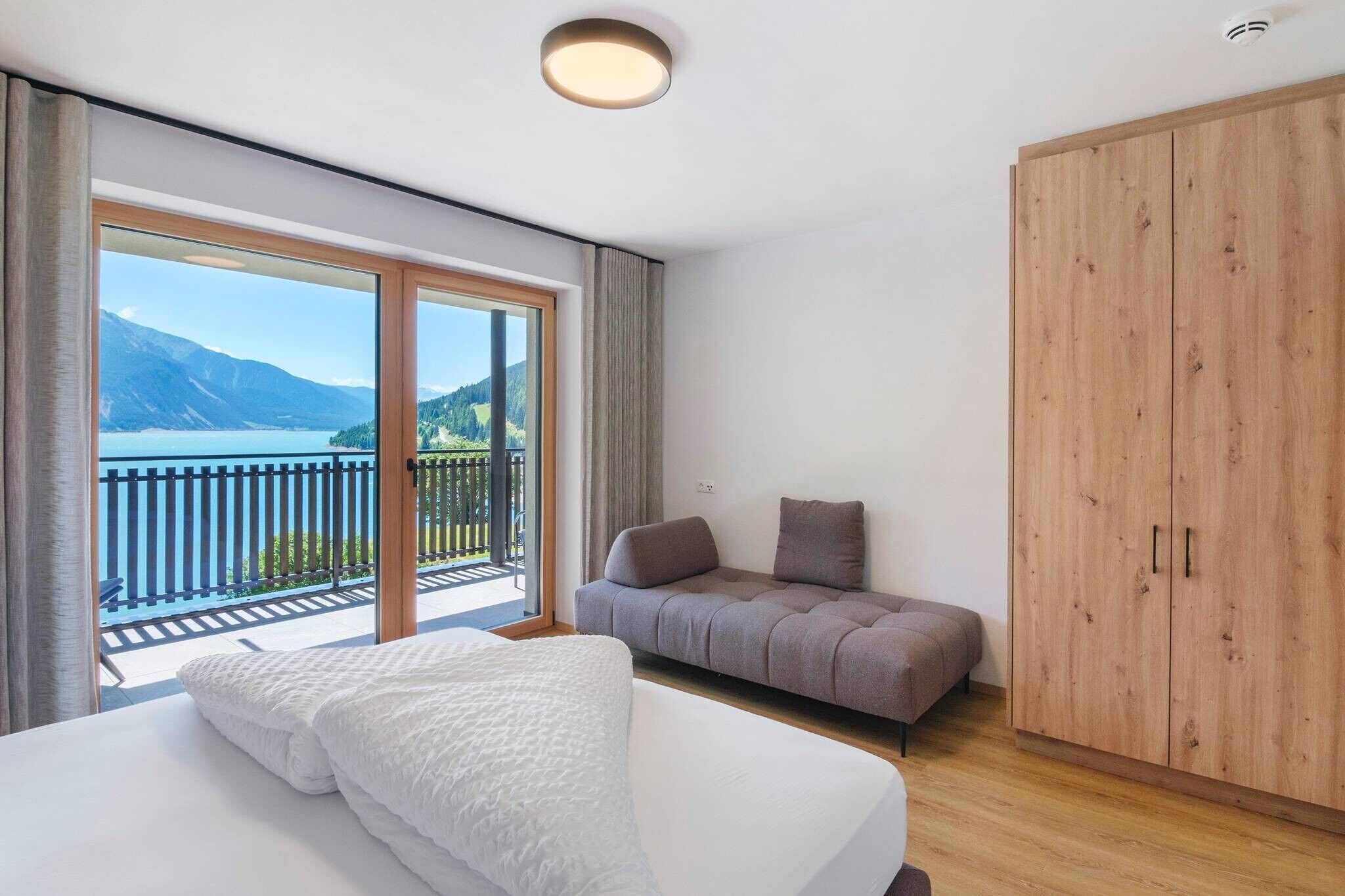 Una camera da letto con un letto, un divano e un armadio. Presente un balcone con vista su montagne e un lago.