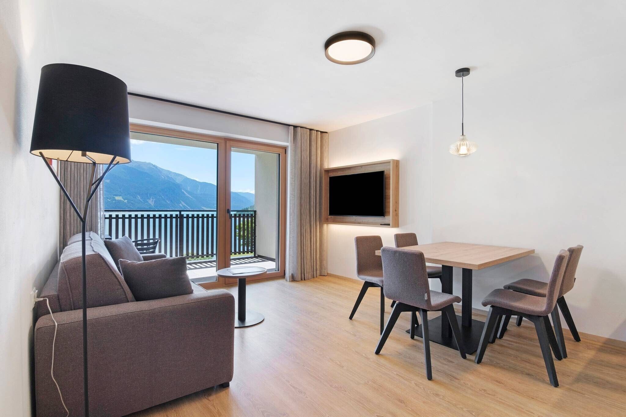 Un soggiorno moderno con un divano, un tavolo da pranzo con sedie, una TV a parete e un balcone con vista sulle montagne.
