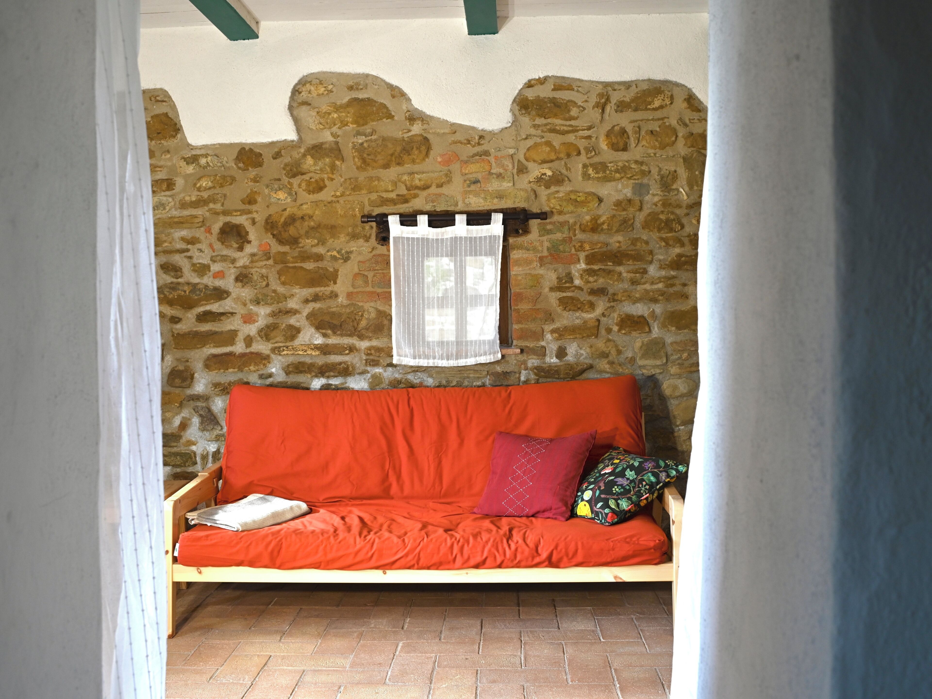 Una habitación con paredes de piedra, un sofá de madera, una ventana con cortina blanca y piso de baldosas.