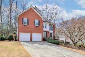 A two-story brick house with a garage, surrounded by trees and a well-maintained lawn. - Spacious 4BR Near Lassiter | Game Room & Basement (Marietta)