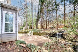 A house with a stone waterfall, a patio with a chair, and a forested area. - Spacious 4BR Near Lassiter | Game Room & Basement (Marietta)