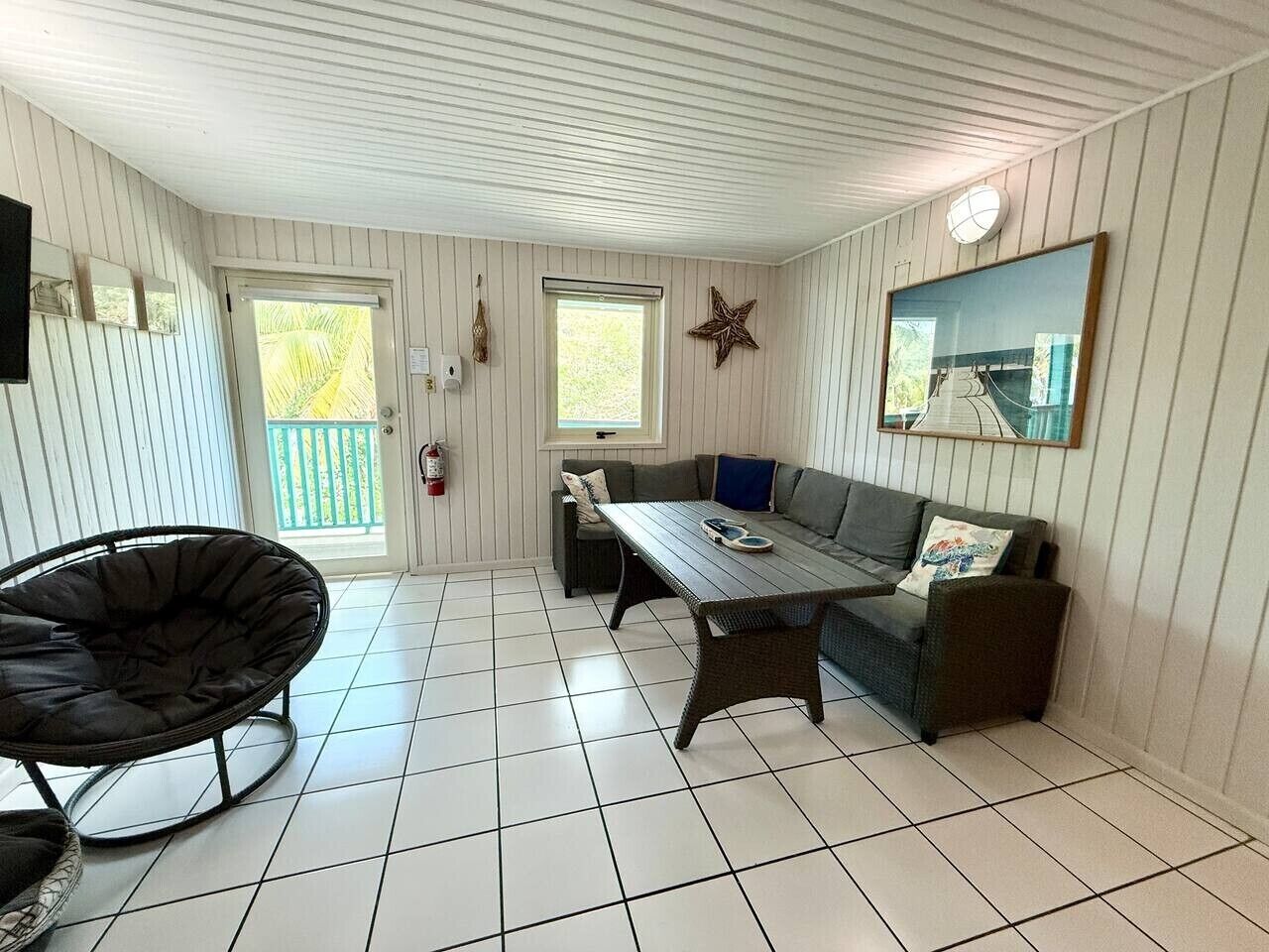Ein Zimmer mit Esstisch, einer Couch und einem Sessel, mit gefliestem Boden und weißen Wänden.