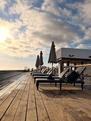 Eine Holzterrasse mit Liegestühlen und Sonnenschirmen mit Blick auf ein Gewässer.