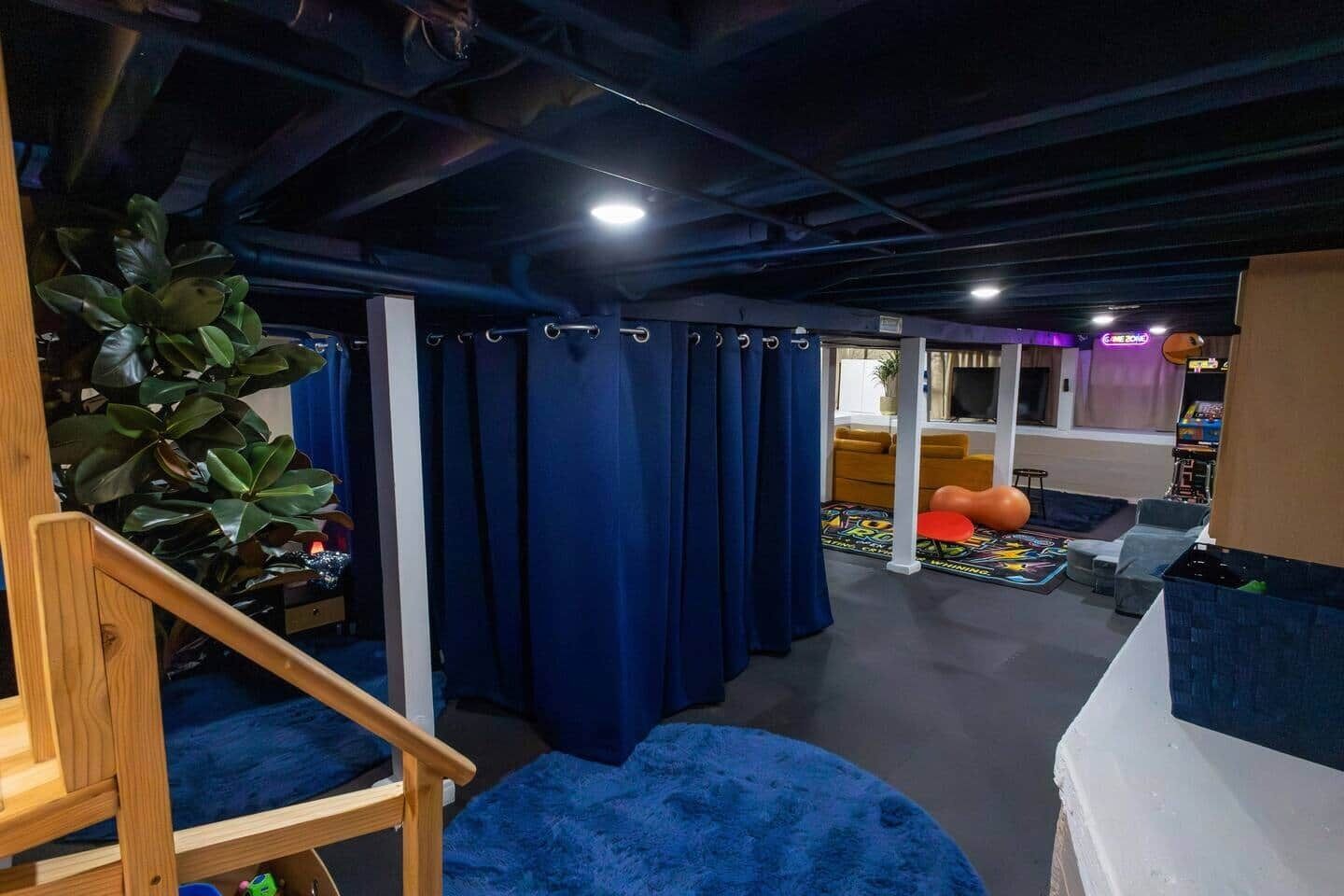 A room with a staircase, blue curtains, a sofa, a gaming area with a TV, and a plant.