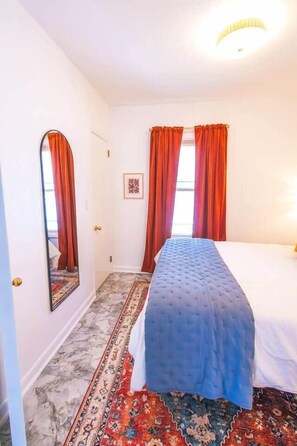 Une chambre à coucher avec un lit, un miroir, une porte et une fenêtre avec des rideaux.