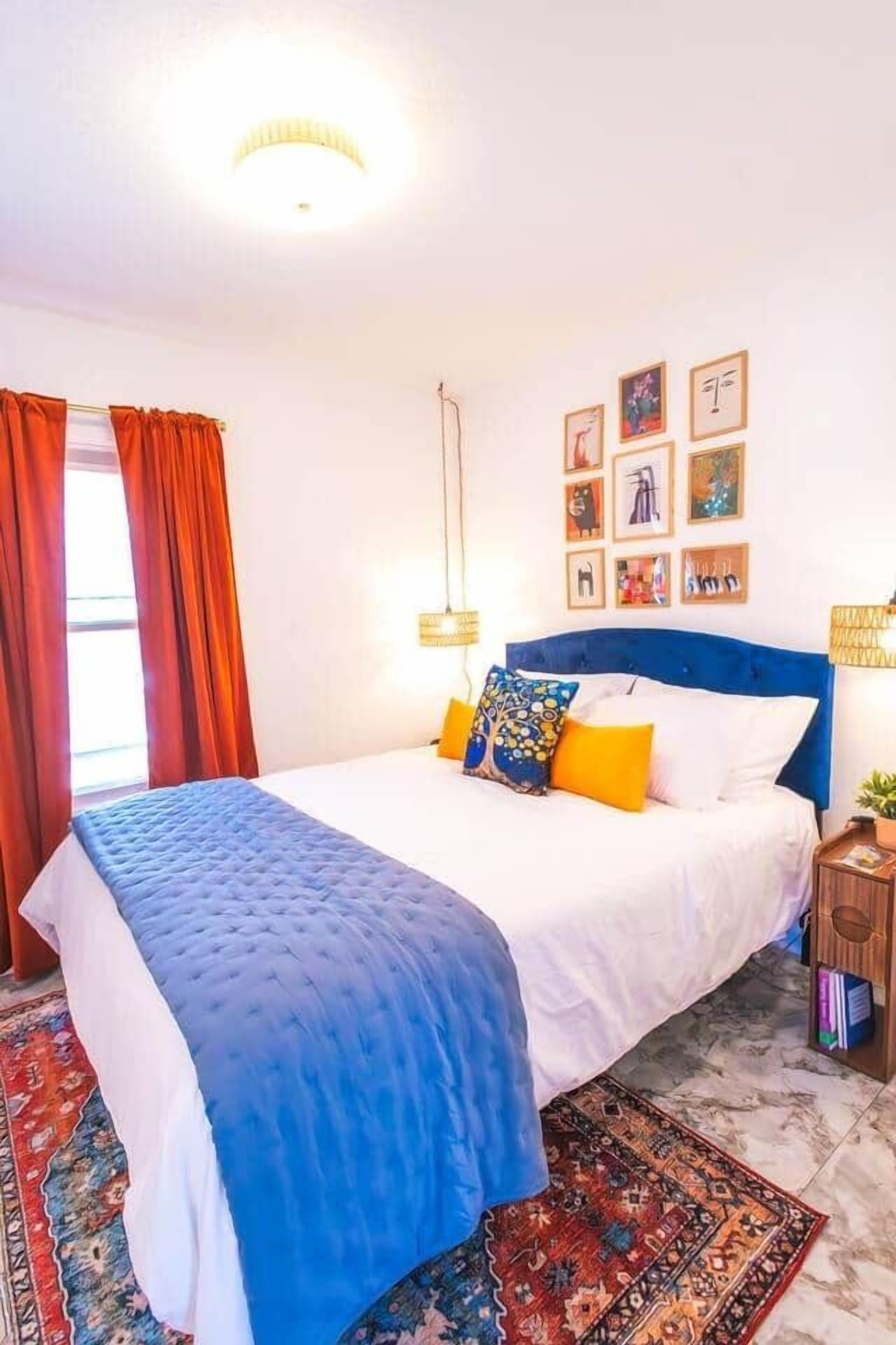 Une chambre à coucher comprenant un lit, une table de chevet, une lampe, une fenêtre avec des rideaux et des œuvres d’art murales.