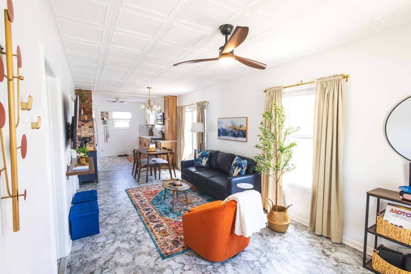 A living room with a sofa, coffee table, and a plant. There is a dining area with a table and chairs. The room has a ceiling fan and a window with curtains.