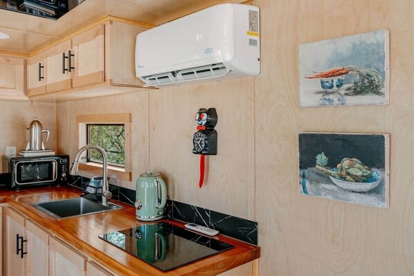 A kitchen with wooden cabinets, a microwave, a kettle, a sink, and a wall-mounted air conditioner.