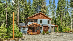 A two-story wooden cabin with a porch, surrounded by a forest.