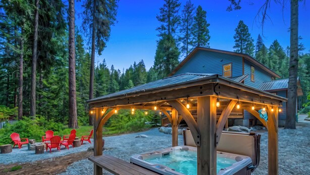 A wooden gazebo with string lights, a hot tub, and red chairs in a forested area.