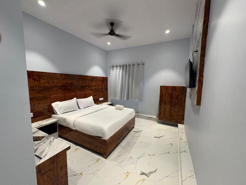 A bedroom with a wooden headboard, a bed with white bedding, a nightstand, a TV mounted on the wall, and a ceiling fan.