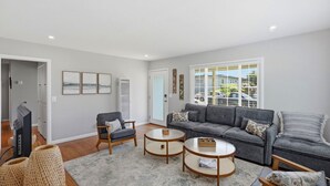 Een woonkamer met een grijze bank, twee fauteuils, een salontafel, een televisie en een groot raam met jaloezieën.