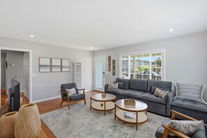 Ein Wohnzimmer mit grauem Sofa, zwei Sesseln, einem Couchtisch, einem Fernseher und einem großen Fenster mit Jalousien.