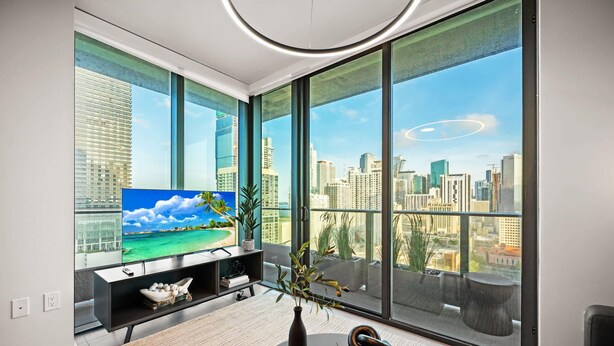 A modern living room with a large TV, a black entertainment unit, and a large window with a city view. - Luxury 2BR/2BA • Rooftop Pool • Downtown Miami (Miami)
