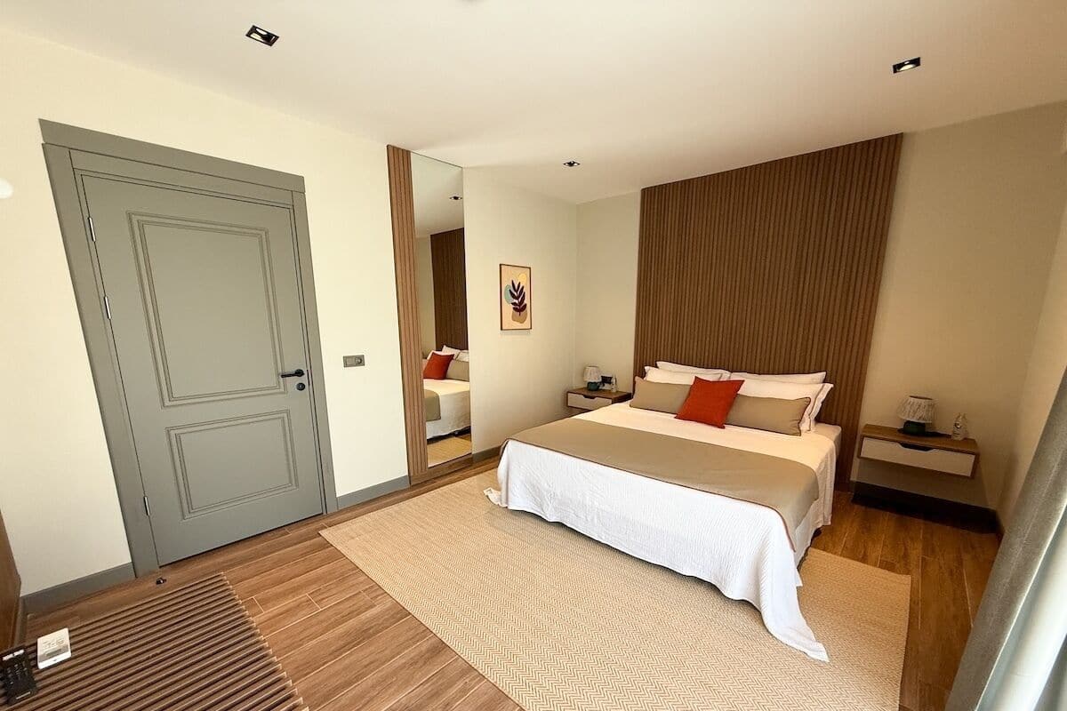 Un dormitorio con cama, mesitas de noche, un espejo, una puerta y suelo de madera.