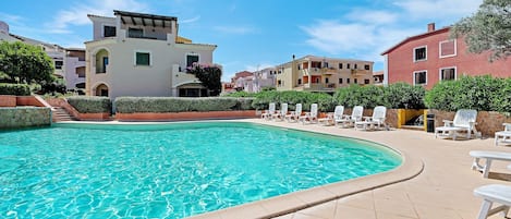 A swimming pool with a tiled deck, lounge chairs, and a building with balconies.