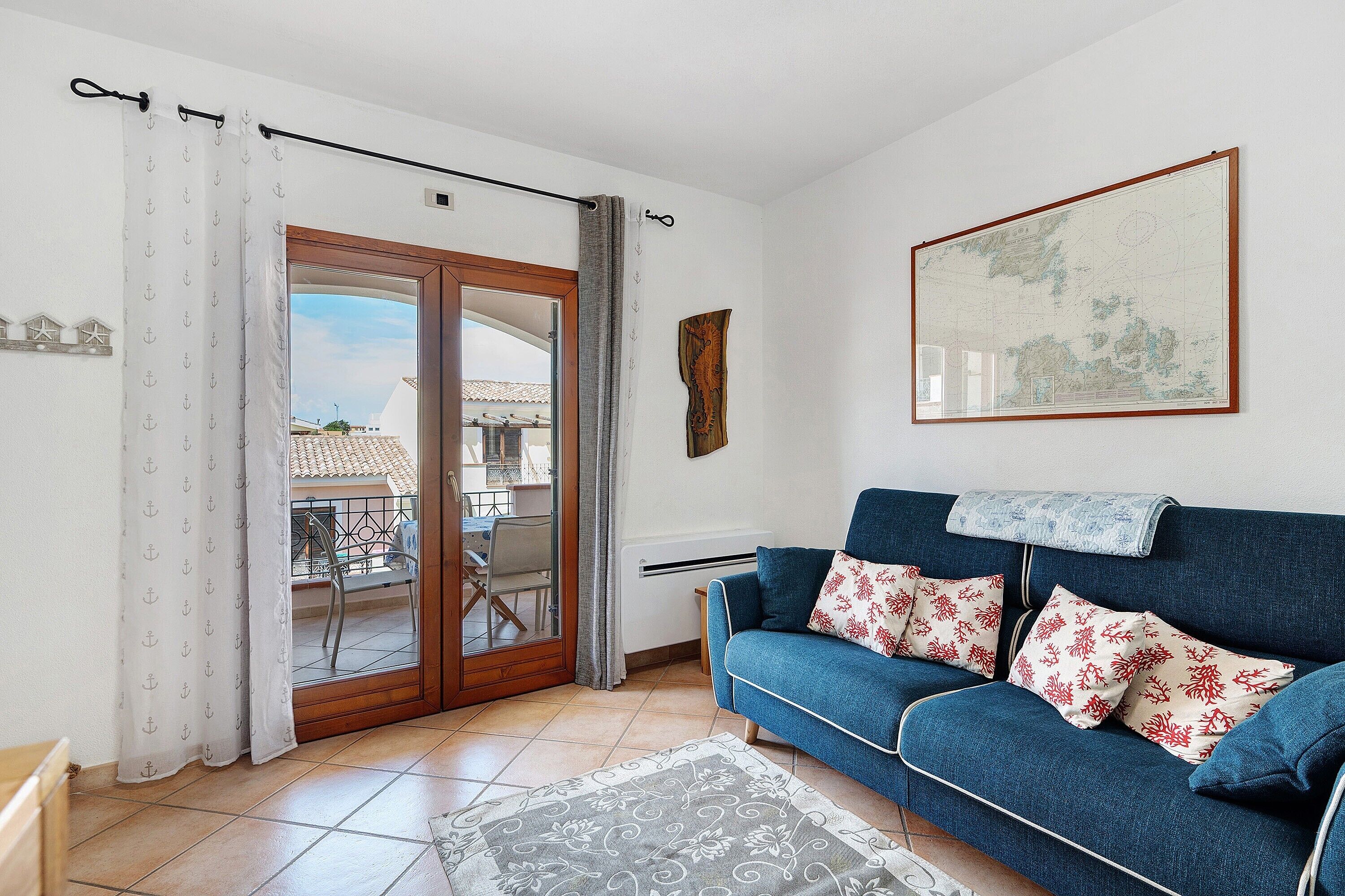 Un salon avec un canapé bleu, des coussins à motifs, une carte accrochée au mur et une vue sur l’extérieur, visible à travers une porte vitrée.