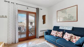 Un salon avec un canapé bleu, des coussins à motifs, une carte accrochée au mur et une vue sur l’extérieur, visible à travers une porte vitrée.