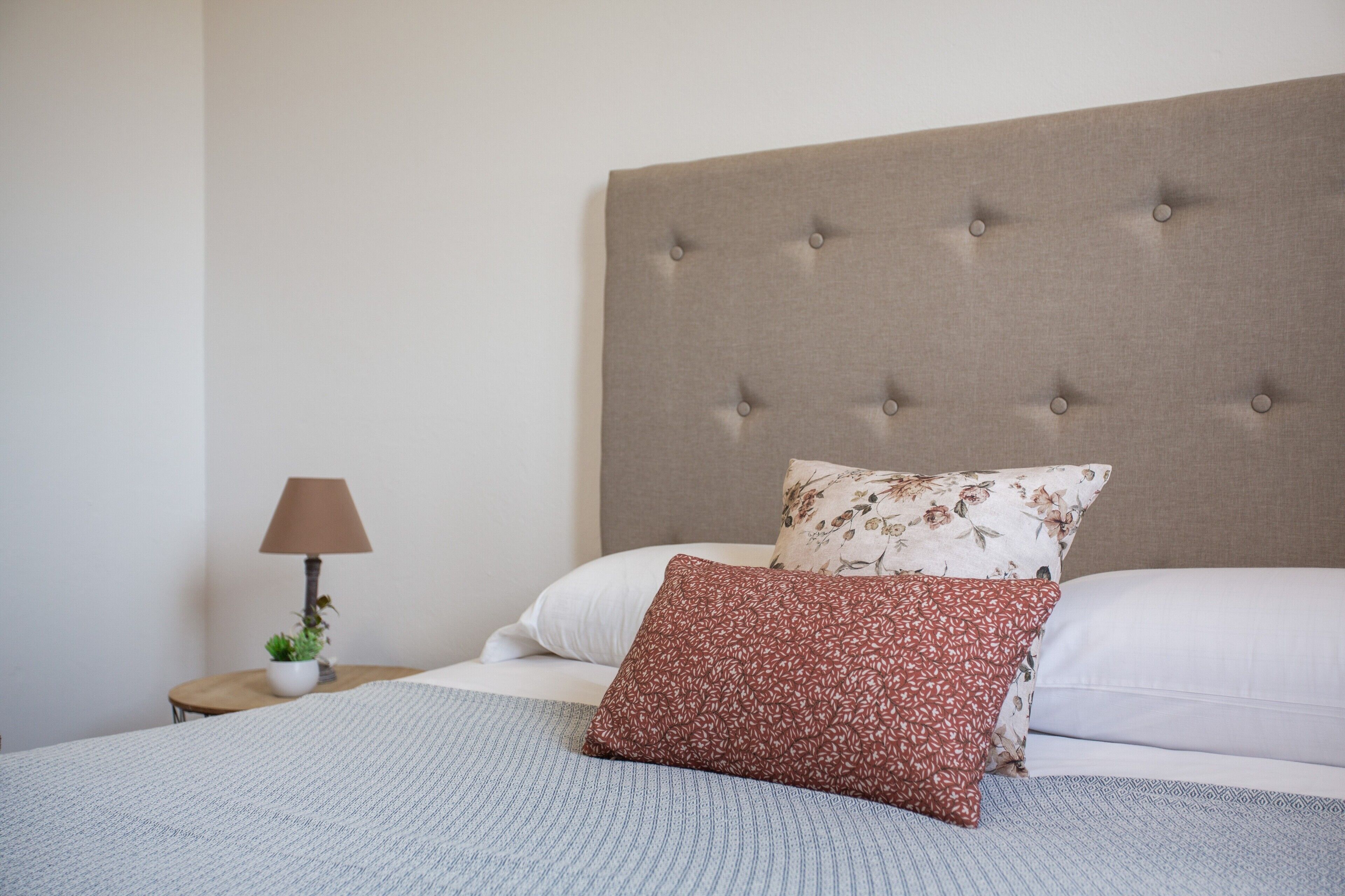 A neatly made bed with a tufted headboard, a bedside table with a lamp, and a small potted plant.