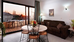 Ein Wohnzimmer mit Sofa, einem Esstisch mit Stühlen, einem Balkon mit Sonnenuntergangsblick und einer Topfpflanze.