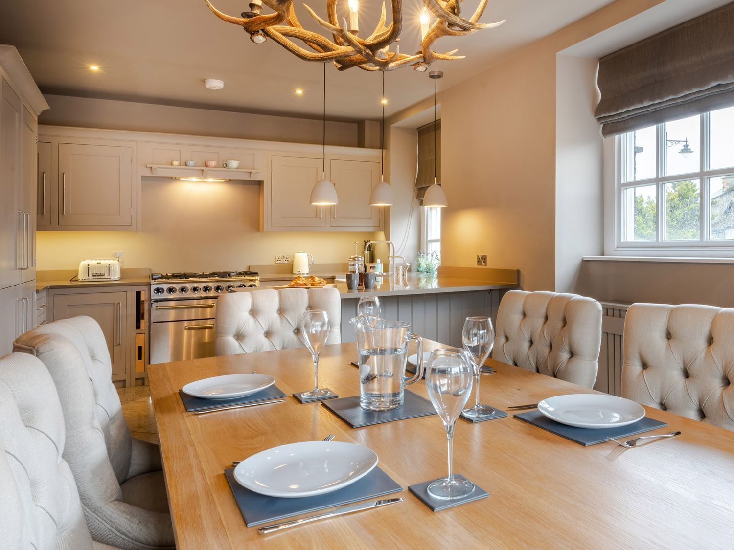A modern kitchen with a dining area, featuring a wooden table set for six, a chandelier, and built-in appliances.