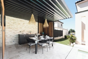 Eine Terrasse im Freien mit Esstisch, Stühlen, einer integrierten Küchenzeile und hängenden Lampen.