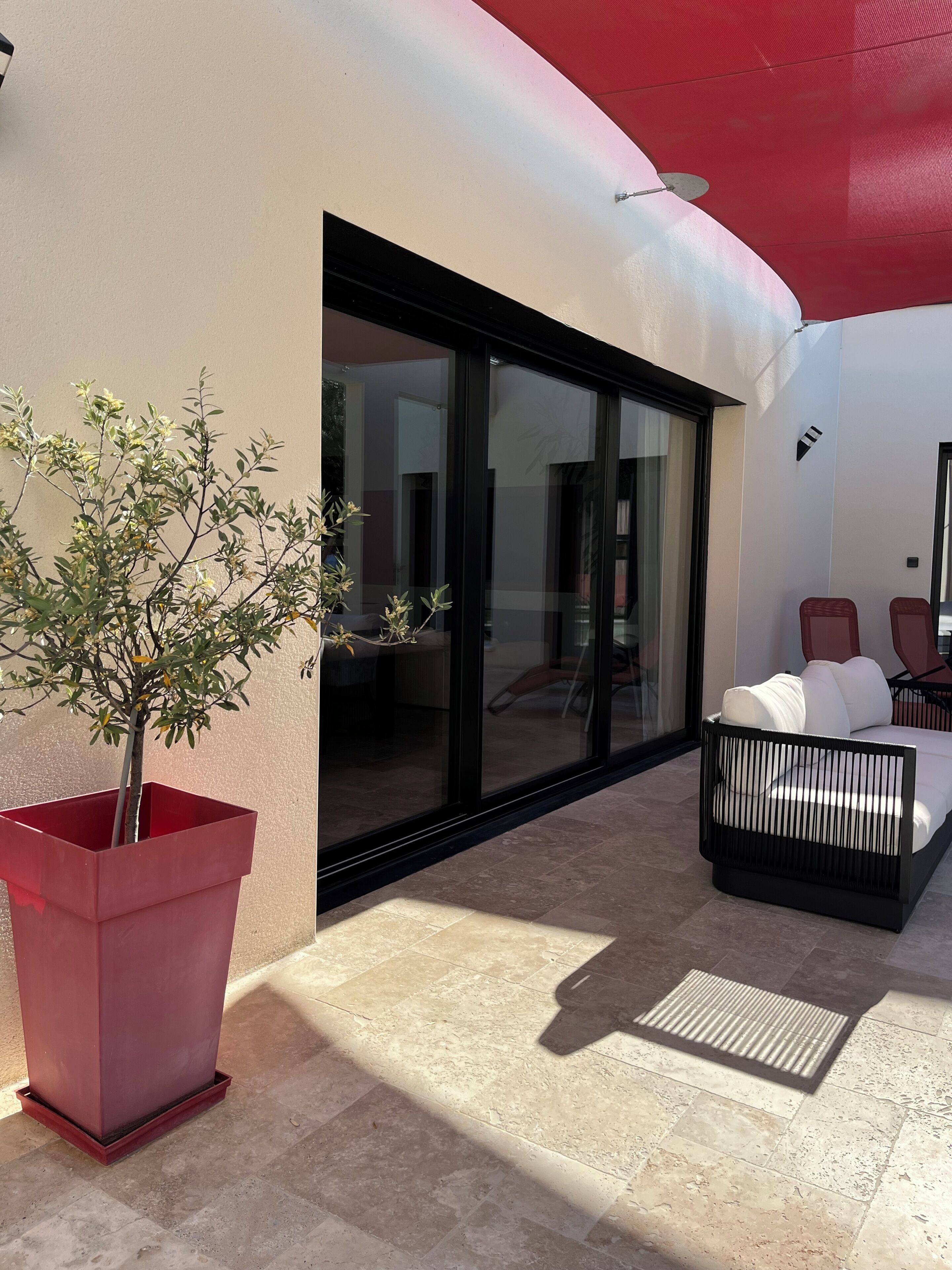 A modern outdoor seating area with a potted plant, a glass door leading to a room, and a red awning.